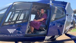 Dos personas se sientan dentro de un helicóptero azul con la puerta abierta, estacionado en una zona cubierta de hierba con vistas al impresionante paisaje durante su "Vuelo en helicóptero por la Costa del Sol (Marbella). - ForTwo - Planes para parejas