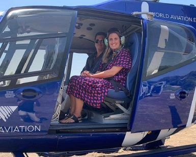 Dos personas se sientan dentro de un helicóptero azul con la puerta abierta, estacionado en una zona cubierta de hierba con vistas al impresionante paisaje durante su "Vuelo en helicóptero por la Costa del Sol (Marbella). - ForTwo - Planes para parejas