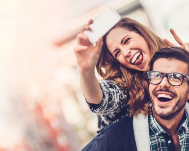 Una mujer sonriente lleva a cuestas a su pareja, ambos haciendo signos de paz para una selfie durante su Viaje sorpresa en pareja por Europa con Waynabox. - ForTwo - Planes para parejas
