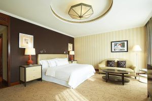 Explora la elegancia de una habitación de Hotel The Westin (Valencia), con una cama grande, pared de acento oscuro, sofá beige, mesa redonda e iluminación suave. - ForTwo - Planes para parejas