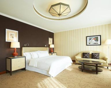 Explora la elegancia de una habitación de Hotel The Westin (Valencia), con una cama grande, pared de acento oscuro, sofá beige, mesa redonda e iluminación suave. - ForTwo - Planes para parejas