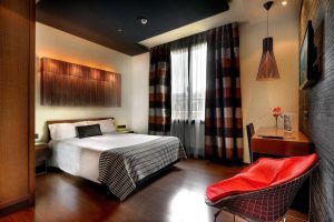 Experimente la comodidad moderna de una habitación del Wilson Boutique Hotel en Barcelona, con cama doble, silla roja, escritorio y cortinas a rayas. La cálida iluminación realza el ambiente acogedor, creando el refugio perfecto tras explorar la vibrante ciudad. - ForTwo - Planes para parejas