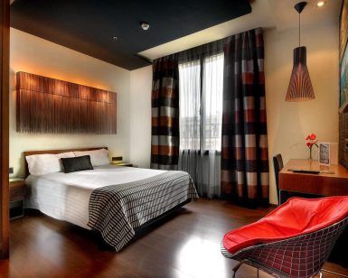 Experimente la comodidad moderna de una habitación del Wilson Boutique Hotel en Barcelona, con cama doble, silla roja, escritorio y cortinas a rayas. La cálida iluminación realza el ambiente acogedor, creando el refugio perfecto tras explorar la vibrante ciudad. - ForTwo - Planes para parejas