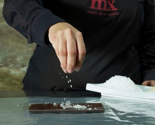 Una persona espolvorea sal sobre un bloque de chocolate rectangular sobre una superficie de aluminio, abrazando verdaderamente la esencia de "Descubre la Xocoexperience en el Museu de la Xocolata y podréis convertiros en maestros del chocolate. - ForTwo - Planes para parejas