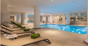 Relájese en tumbonas con toallas verdes en la piscina cubierta del Meliá Madrid Princesa, rodeado de máquinas de gimnasio. Disfrute de la rejuvenecedora Hidroterapia en el Spa Yhi Wellness del hotel Meliá Madrid Princesa 5 estrellas para una experiencia revitalizante. - ForTwo - Planes para parejas