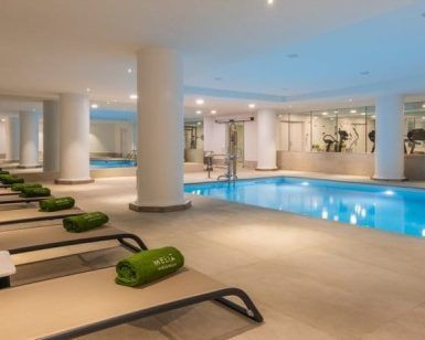 Relájese en tumbonas con toallas verdes en la piscina cubierta del Meliá Madrid Princesa, rodeado de máquinas de gimnasio. Disfrute de la rejuvenecedora Hidroterapia en el Spa Yhi Wellness del hotel Meliá Madrid Princesa 5 estrellas para una experiencia revitalizante. - ForTwo - Planes para parejas