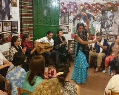 En una sala pequeña y abarrotada, una mujer con un vestido azul interpreta apasionadamente flamenco para un público embelesado, que recuerda al cautivador Espectáculo de flamenco en la Zambra María la Canastera. - ForTwo - Planes para parejas
