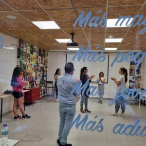 En un aula vibrante con carteles coloridos, los participantes se involucran en una actividad enérgica como Clases de Sevillanas en Granada: domina el compás y disfruta del folclore, mientras las palabras resuenan suavemente en la mampara de cristal, enriqueciendo el ambiente con el espíritu folclórico de Granada. - ForTwo - Planes para parejas