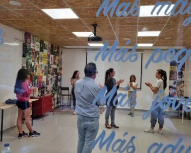 En un aula vibrante con carteles coloridos, los participantes se involucran en una actividad enérgica como Clases de Sevillanas en Granada: domina el compás y disfruta del folclore, mientras las palabras resuenan suavemente en la mampara de cristal, enriqueciendo el ambiente con el espíritu folclórico de Granada. - ForTwo - Planes para parejas