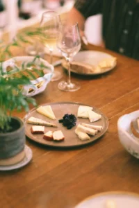 Una mesa de madera con una tabla de quesos y copas de vino invita a disfrutar de la "Cata de 5 Vinos Naturales y 5 Quesos: Una Experiencia para Amantes del Buen Sabor". Una planta en maceta añade un toque natural a la izquierda. - ForTwo - Planes para parejas