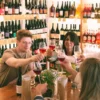 En una acogedora tienda de vinos, la gente disfrutó de la "Cata de 5 Vinos Naturales y 5 Quesos: Una Experiencia para Amantes del Buen Sabor", alzando copas de vinos naturales en medio de estantes llenos de botellas. - ForTwo - Planes para parejas