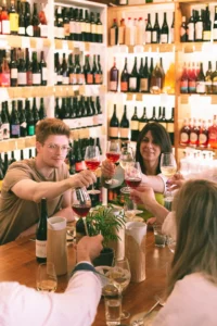 En una acogedora tienda de vinos, la gente disfrutó de la "Cata de 5 Vinos Naturales y 5 Quesos: Una Experiencia para Amantes del Buen Sabor", alzando copas de vinos naturales en medio de estantes llenos de botellas. - ForTwo - Planes para parejas