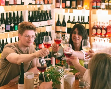 En una acogedora tienda de vinos, la gente disfrutó de la "Cata de 5 Vinos Naturales y 5 Quesos: Una Experiencia para Amantes del Buen Sabor", alzando copas de vinos naturales en medio de estantes llenos de botellas. - ForTwo - Planes para parejas