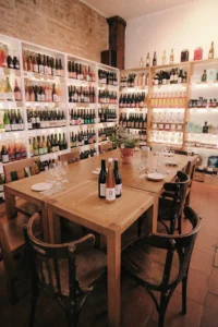 Una acogedora tienda con mesa y sillas de madera que ofrece "Cata de 5 Vinos Naturales y 5 Quesos: Una Experiencia para Amantes del Buen Sabor", con vinos naturales elegantemente expuestos en los estantes. - ForTwo - Planes para parejas