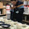 Inmerso en una mesa de cafés de especialidad y abastos, una persona saborea el aroma de su taza, experimentando la esencia de una Cata de cafés de especialidad. - ForTwo - Planes para parejas