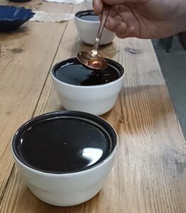Con elegancia, una persona sostiene una cuchara sobre cuencos de líquido oscuro, listos para la Cata de cafés de especialidad en una mesa de madera rústica. - ForTwo - Planes para parejas