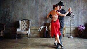 En una sala con poca luz, adornada con flores y decoración vintage, una pareja baila tango con gracia, capturando la pasión de las famosas Clases de Baile en Sevilla de Sevilla: Flamenco, Ballet, Sevillanas, Bailes Latinos y de Salón. - ForTwo - Planes para parejas