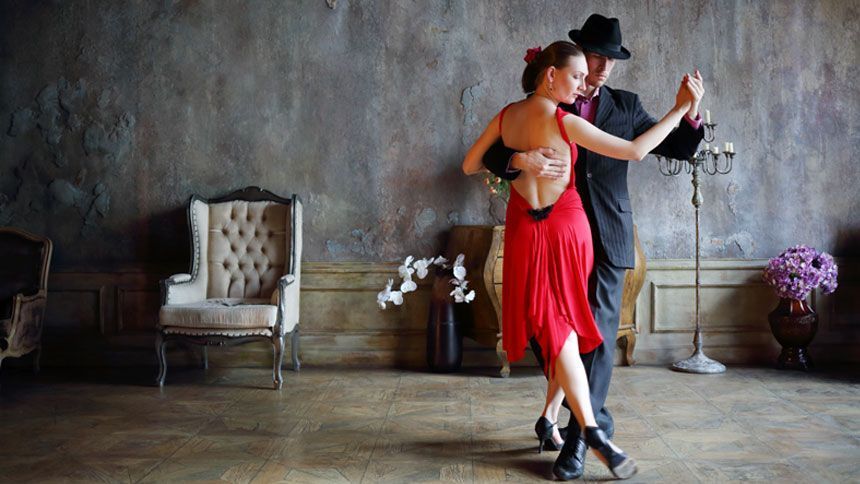 En una sala con poca luz, adornada con flores y decoración vintage, una pareja baila tango con gracia, capturando la pasión de las famosas Clases de Baile en Sevilla de Sevilla: Flamenco, Ballet, Sevillanas, Bailes Latinos y de Salón. - ForTwo - Planes para parejas