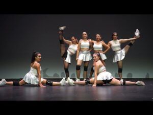 Seis bailarines vestidos de blanco realizan poses dinámicas y splits en el escenario de Sevilla, cautivando al público con los ritmos de "Clases de Salsa en Sevilla: Descubre el Ritmo Latino". - ForTwo - Planes para parejas