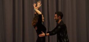 Una pareja baila con gracia al estilo de "Clases de Salsa en Sevilla: Descubre el Ritmo Latino", con la mujer levantando los brazos y el hombre sujetándola por la cintura, todo ello frente a un fondo de cortinas. - ForTwo - Planes para parejas