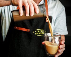 Una persona de Coffee Hackers: Vive una Cata Sensorial de Café como Nunca Antes vierte café de una coctelera a un vaso, y su delantal negro muestra el icónico logotipo. - ForTwo - Planes para parejas