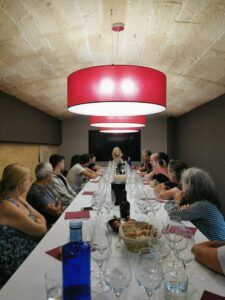 Los asistentes al Curso de Cata de Vinos en Barcelona, sentados alrededor de una larga mesa con copas de vino, escuchan atentamente a un experto al frente mientras las luces rojas del techo crean un ambiente cálido. - ForTwo - Planes para parejas