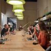 En el corazón de Barcelona, bajo grandes lámparas verdes, la gente se reúne en una larga mesa en una tienda de vinos para un improvisado Curso de Cata de Vinos en Barcelona, disfrutando de vinos e historias que fluyen libremente. - ForTwo - Planes para parejas
