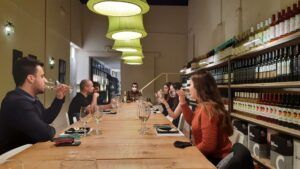 En el corazón de Barcelona, bajo grandes lámparas verdes, la gente se reúne en una larga mesa en una tienda de vinos para un improvisado Curso de Cata de Vinos en Barcelona, disfrutando de vinos e historias que fluyen libremente. - ForTwo - Planes para parejas