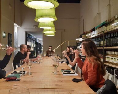 En el corazón de Barcelona, bajo grandes lámparas verdes, la gente se reúne en una larga mesa en una tienda de vinos para un improvisado Curso de Cata de Vinos en Barcelona, disfrutando de vinos e historias que fluyen libremente. - ForTwo - Planes para parejas