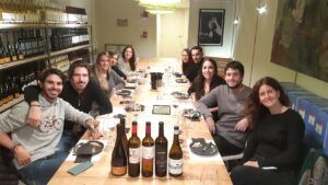 Un grupo animado se reúne en una mesa de comedor, rodeado de botellas de vino y copas, mientras la acogedora sala vibra de entusiasmo por el Curso de Cata de Vinos en Barcelona. - ForTwo - Planes para parejas