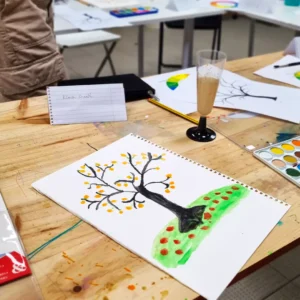 Esta encantadora pintura de un árbol otoñal adorna una mesa entre materiales de arte y una bebida, creando el ambiente perfecto para principiantes en el "Curso de Iniciación a la Acuarela en Granada: libera tu creatividad artística. - ForTwo - Planes para parejas