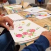Una persona pinta cuidadosamente flores vibrantes con acuarelas en un cuaderno de bocetos en una mesa de madera llena de materiales de arte, participando en el "Curso de Iniciación a la Acuarela en Granada: libera tu creatividad artística". - ForTwo - Planes para parejas