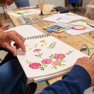 Una persona pinta cuidadosamente flores vibrantes con acuarelas en un cuaderno de bocetos en una mesa de madera llena de materiales de arte, participando en el "Curso de Iniciación a la Acuarela en Granada: libera tu creatividad artística". - ForTwo - Planes para parejas