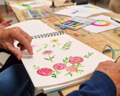 Una persona pinta cuidadosamente flores vibrantes con acuarelas en un cuaderno de bocetos en una mesa de madera llena de materiales de arte, participando en el "Curso de Iniciación a la Acuarela en Granada: libera tu creatividad artística". - ForTwo - Planes para parejas