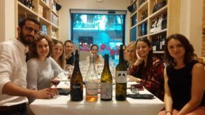 Un grupo de personas sonriendo alrededor de una mesa con vino en un acogedor restaurante de Barcelona, disfrutando del Curso de Maridaje de Vinos en Barcelona: Quesos, Ibéricos o Chocolate. - ForTwo - Planes para parejas
