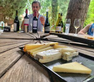 Una tabla de quesos y botellas de vino sobre una mesa de madera al aire libre capturan la esencia de Barcelona, resaltando el Curso de Maridaje de Vinos en Barcelona: Quesos, Ibéricos o Chocolate mientras alguien al fondo disfruta de esta combinación perfecta en medio de un entorno iluminado por el sol. - ForTwo - Planes para parejas