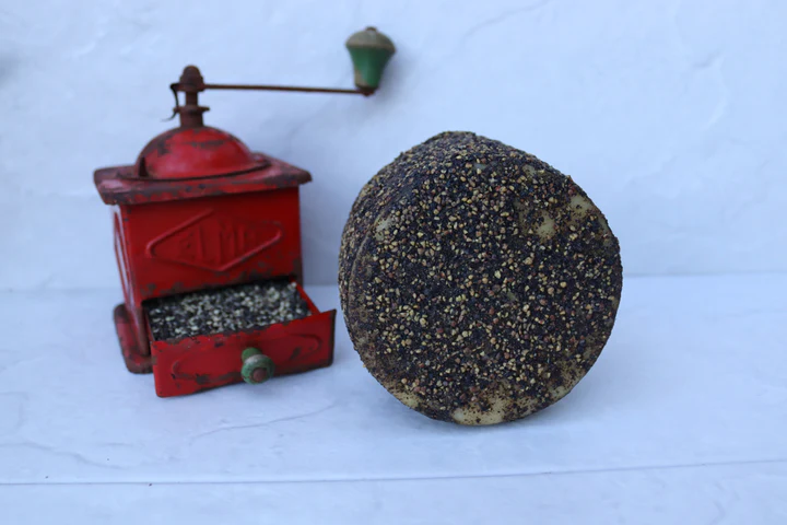 Un queso redondo de Degustación de quesos artesanos en Quesería Cortijo El Aserradero (Granada), recubierto de granos de pimienta, complementa un molinillo de café rojo vintage sobre un fondo blanco, resaltando el encanto artesanal de los quesos granadinos. - ForTwo - Planes para parejas