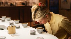 Una mujer con camisa amarilla disfruta de una degustación de café, saboreando los aromas de los productos de "Cata de Café Abierta en Barcelona: ¡Descubre los Secretos del Café con Cafés el Magnífico!" sobre una mesa blanca en el interior. - ForTwo - Planes para parejas