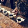 En el "Taller de Café" de The Coffee Hackers en Barcelona, un experto en café utiliza una cuchara para degustar muestras en una mesa de madera, explorando varias tazas alineadas para esta experiencia sensorial. - ForTwo - Planes para parejas