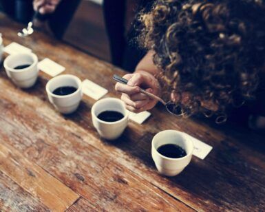 En el "Taller de Café" de The Coffee Hackers en Barcelona, un experto en café utiliza una cuchara para degustar muestras en una mesa de madera, explorando varias tazas alineadas para esta experiencia sensorial. - ForTwo - Planes para parejas