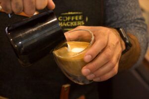 En un acogedor café de Barcelona, un barista del Taller de Café en The Coffee Hackers vierte leche en una taza de café, creando con maestría el arte del latte, un sello distintivo entre los locales. - ForTwo - Planes para parejas
