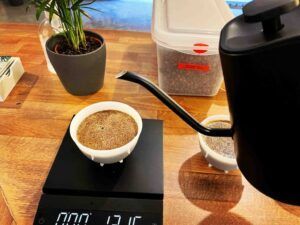 Sirviendo café en una taza blanca sobre una báscula digital en el Taller de Café de The Coffee Hackers Barcelona, con café molido y una planta cerca sobre una mesa de madera. - ForTwo - Planes para parejas