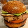 Un primer plano de una hamburguesa con pan con semillas de sésamo, aguacate, tomate y hamburguesa de carne, una verdadera obra maestra del Taller de Comida Rápida Saludable en Sevilla, presenta vasos y una bebida difuminada en el fondo. - ForTwo - Planes para parejas