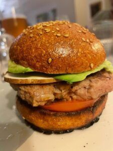 Un primer plano de una hamburguesa con pan con semillas de sésamo, aguacate, tomate y hamburguesa de carne, una verdadera obra maestra del Taller de Comida Rápida Saludable en Sevilla, presenta vasos y una bebida difuminada en el fondo. - ForTwo - Planes para parejas