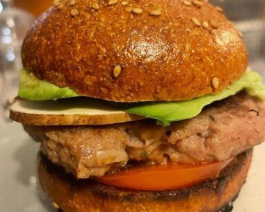 Un primer plano de una hamburguesa con pan con semillas de sésamo, aguacate, tomate y hamburguesa de carne, una verdadera obra maestra del Taller de Comida Rápida Saludable en Sevilla, presenta vasos y una bebida difuminada en el fondo. - ForTwo - Planes para parejas