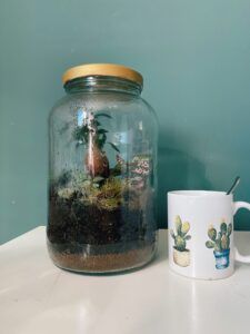Un gran terrario de tarros junto a una taza con temática de cactus sobre una superficie blanca muestra el estilo granadino, inspirado en el taller de jardín eterno en Granada: crea tu propio terrario autosostenible. - ForTwo - Planes para parejas
