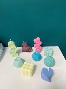 Una gama de velas decorativas coloridas y con formas únicas se exhiben sobre una superficie blanca sobre un fondo verde azulado, destacando el Taller de Velas Decorativas en Granada: crea tus propias velas aromáticas. - ForTwo - Planes para parejas