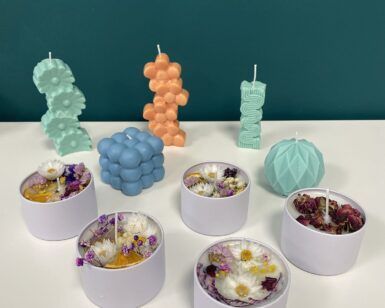 Las velas coloridas y de formas únicas de Taller de Velas Decorativas en Granada adornan hermosamente una mesa blanca contra una pared verde azulado, infundiendo su espacio con la esencia de Granada. - ForTwo - Planes para parejas