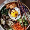 Un colorido tazón de bibimbap con un huevo frito, verduras vibrantes, champiñones y arroz se presenta artísticamente junto con palillos, que recuerdan al Taller de cocina coreana en Sevilla. - ForTwo - Planes para parejas