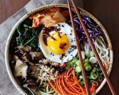 Un colorido tazón de bibimbap con un huevo frito, verduras vibrantes, champiñones y arroz se presenta artísticamente junto con palillos, que recuerdan al Taller de cocina coreana en Sevilla. - ForTwo - Planes para parejas